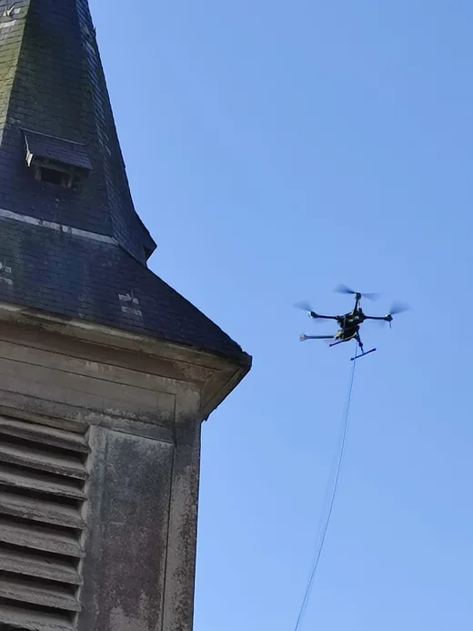 Un drone en vol stationnaire à hauteur de la flèche en ardoise d'un clocher d'église.
