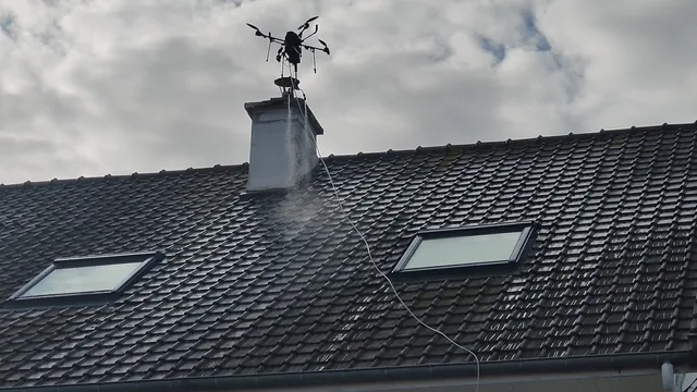 Le drone au-dessus d'une cheminée pulvérise l'anti-mousse sur une toiture sombre.